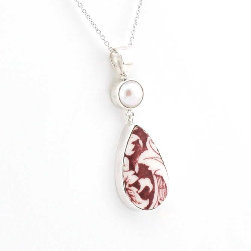 A silver pendant necklace featuring a round white pearl and a teardrop-shaped pendant with a marbled burgundy and white pattern, set against a plain white background.