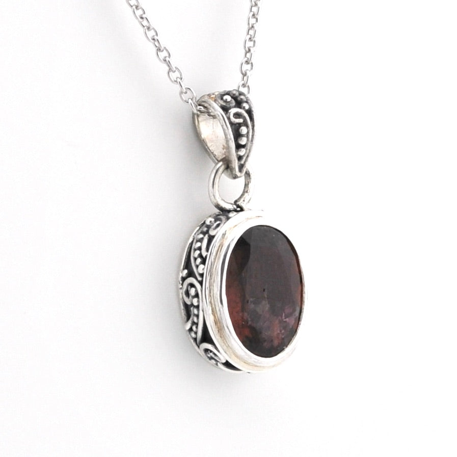 A silver pendant necklace featuring an oval dark brown gemstone in an ornate, textured silver bezel setting, suspended from a silver chain against a plain white background.