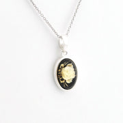 A silver oval pendant necklace featuring a black background with a detailed cream-colored rose and gold accents, suspended from a silver chain against a plain white background.