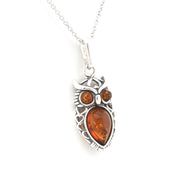 An owl pendant necklace featuring a silver-toned metal frame with a large teardrop-shaped amber gemstone in the center and two smaller round amber gemstones for the eyes, suspended from a silver chain on a white background.