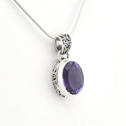 A silver pendant necklace featuring an oval-cut purple amethyst gemstone in a detailed filigree bezel setting, suspended from a silver snake chain against a plain white background.