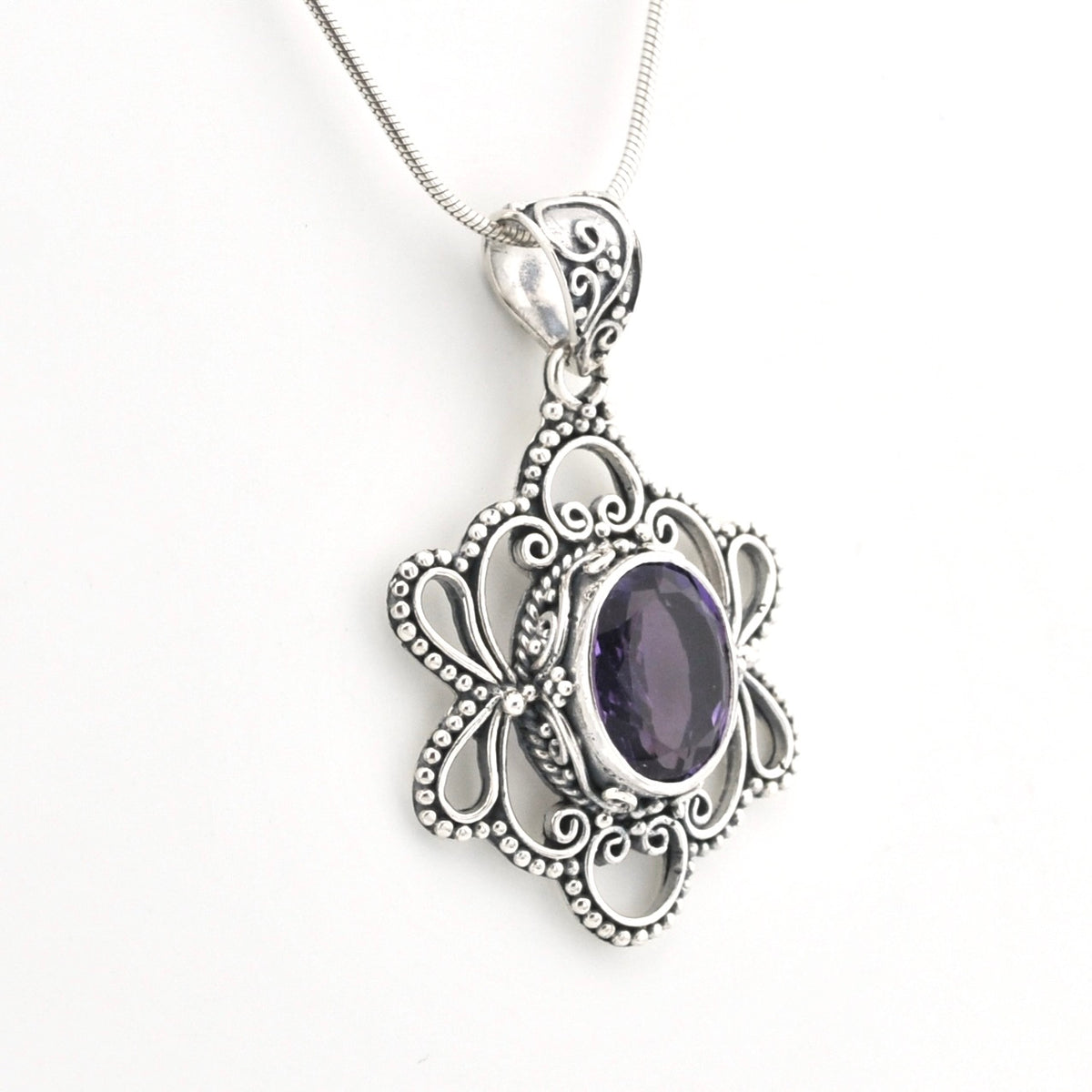 An ornate silver pendant necklace featuring a central oval purple gemstone surrounded by intricate filigree and beaded scrollwork, suspended from a delicate silver snake chain against a plain white background.
