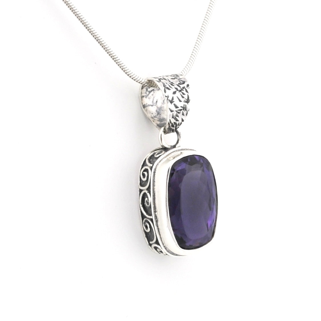 A silver pendant necklace featuring a rectangular purple gemstone with ornate silver filigree detailing on the sides, suspended from a silver snake chain against a plain white background.