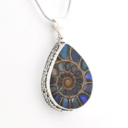 A teardrop-shaped pendant necklace featuring a detailed ammolite fossil inlay with iridescent blue, green, and brown hues, set in a silver-toned filigree border and suspended from a silver chain against a white background.