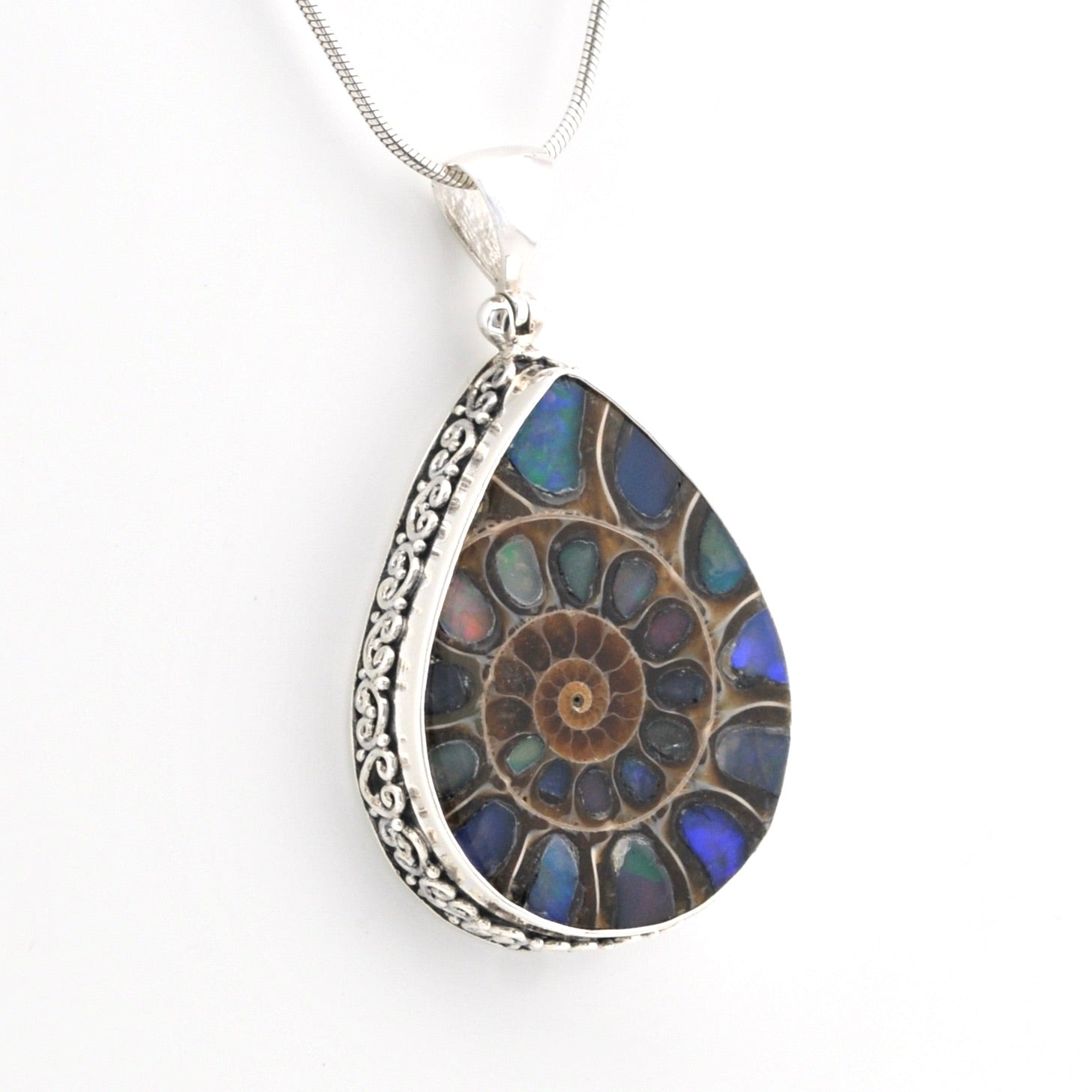 A teardrop-shaped pendant necklace featuring a detailed ammolite fossil inlay with iridescent blue, green, and brown hues, set in a silver-toned filigree border and suspended from a silver chain against a white background.