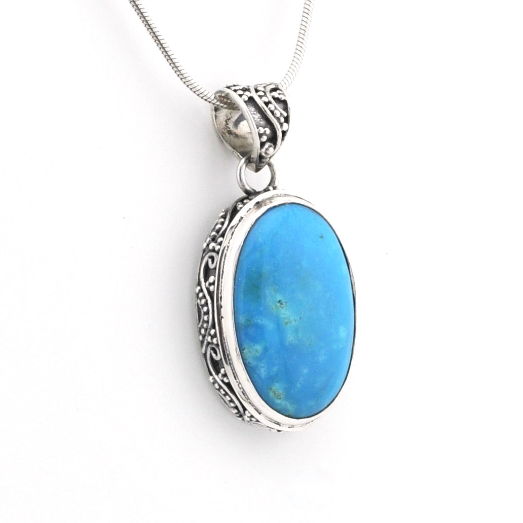 An oval pendant necklace featuring a vibrant blue turquoise stone set in an ornate silver filigree bezel, suspended from a silver snake chain against a plain white background.