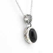 A silver pendant necklace featuring an oval black onyx stone set in an ornate, filigree-style silver bezel, suspended from a delicate silver chain against a plain white background.
