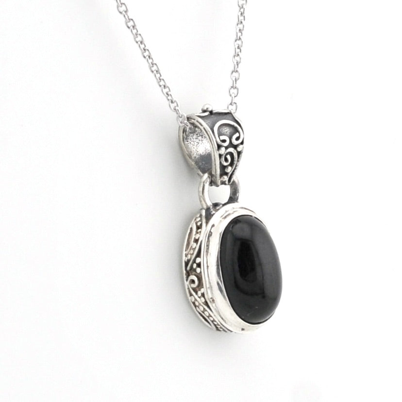 A silver pendant necklace featuring an oval black onyx stone set in an ornate, filigree-style silver bezel, suspended from a delicate silver chain against a plain white background.