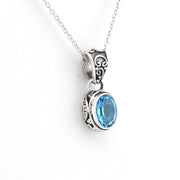A silver pendant necklace featuring an oval-cut light blue gemstone in an ornate, textured silver bezel setting, suspended from a delicate silver chain against a plain white background.