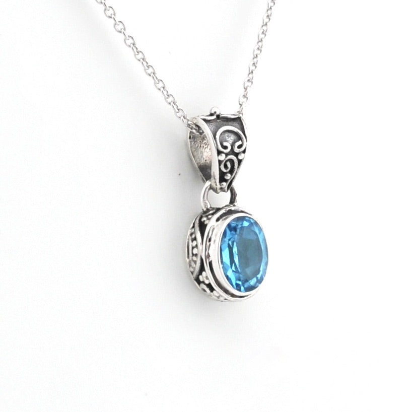 A silver pendant necklace featuring an oval-cut light blue gemstone in an ornate, textured silver bezel setting, suspended from a delicate silver chain against a plain white background.