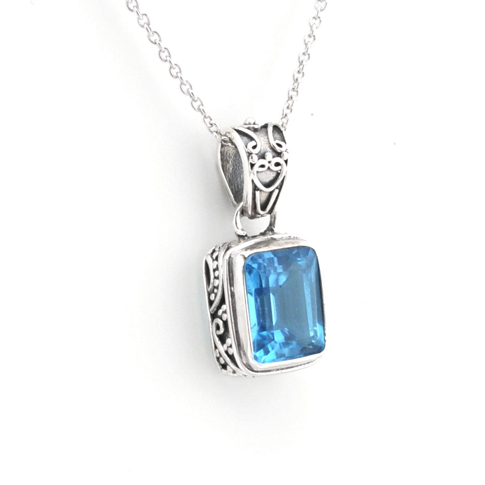 A silver pendant necklace featuring a rectangular blue gemstone in an ornate silver bezel setting, suspended from a delicate silver chain against a plain white background.
