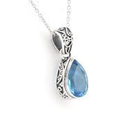 A silver pendant necklace featuring a teardrop-shaped blue gemstone in an ornate, filigree-wrapped setting, suspended from a delicate silver chain against a plain white background.