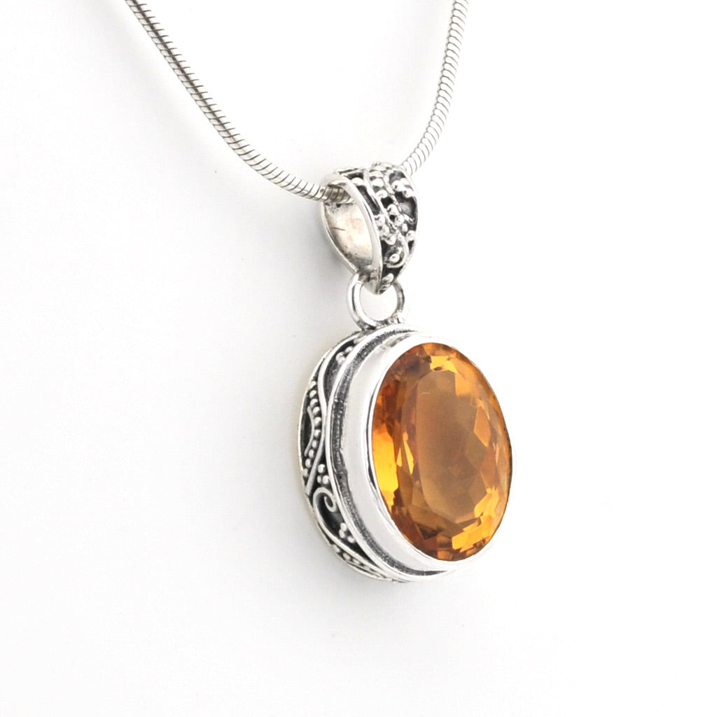 A silver pendant necklace featuring an oval faceted amber-orange gemstone in an ornate silver bezel setting, suspended from a silver snake chain against a plain white background.