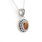 A silver-toned oval pendant necklace featuring a central orange gemstone, detailed with textured rope borders and a filigree bail, shown on a white background.