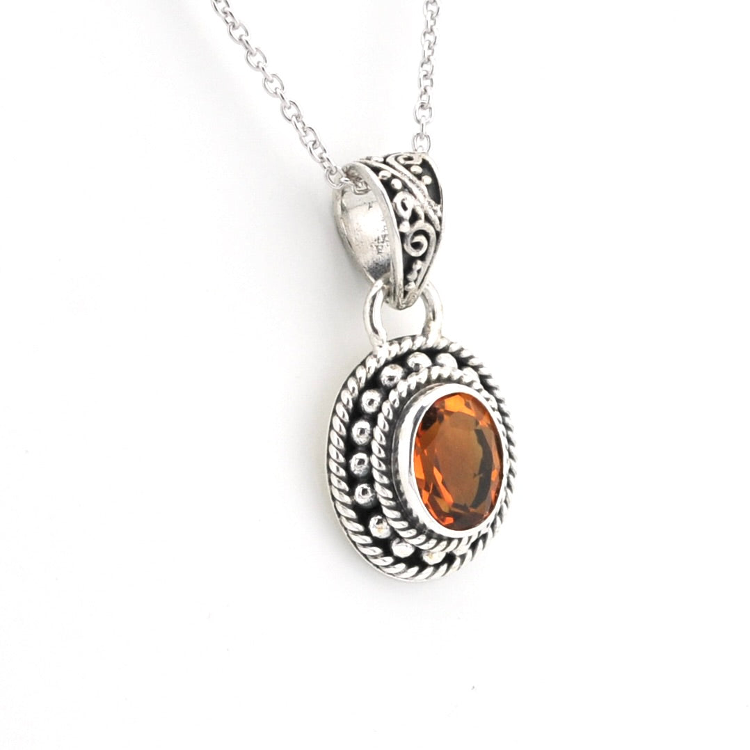 A silver-toned oval pendant necklace featuring a central orange gemstone, detailed with textured rope borders and a filigree bail, shown on a white background.
