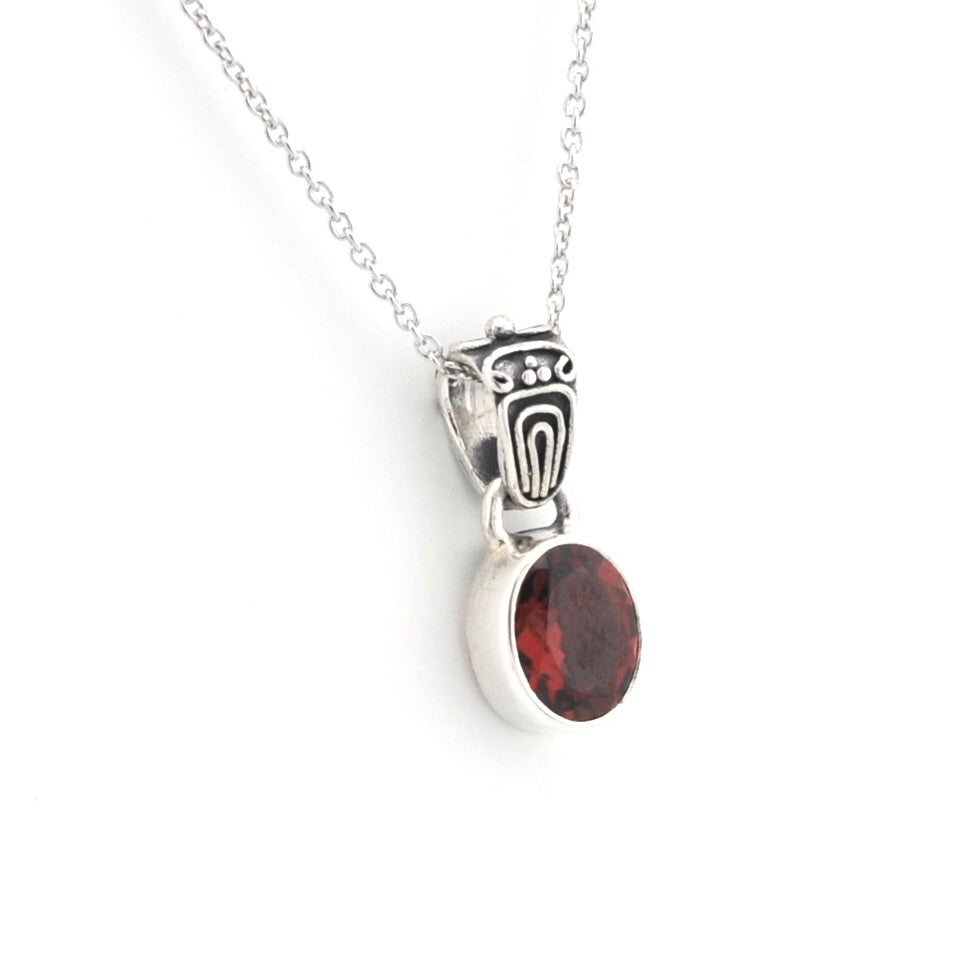 A silver pendant necklace featuring a round, faceted dark red gemstone in a bezel setting, suspended from an ornate silver bail on a delicate chain, set against a plain white background.