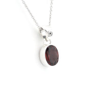 A silver pendant necklace featuring an oval dark red garnet stone in a polished bezel setting, suspended from a delicate silver chain against a plain white background.