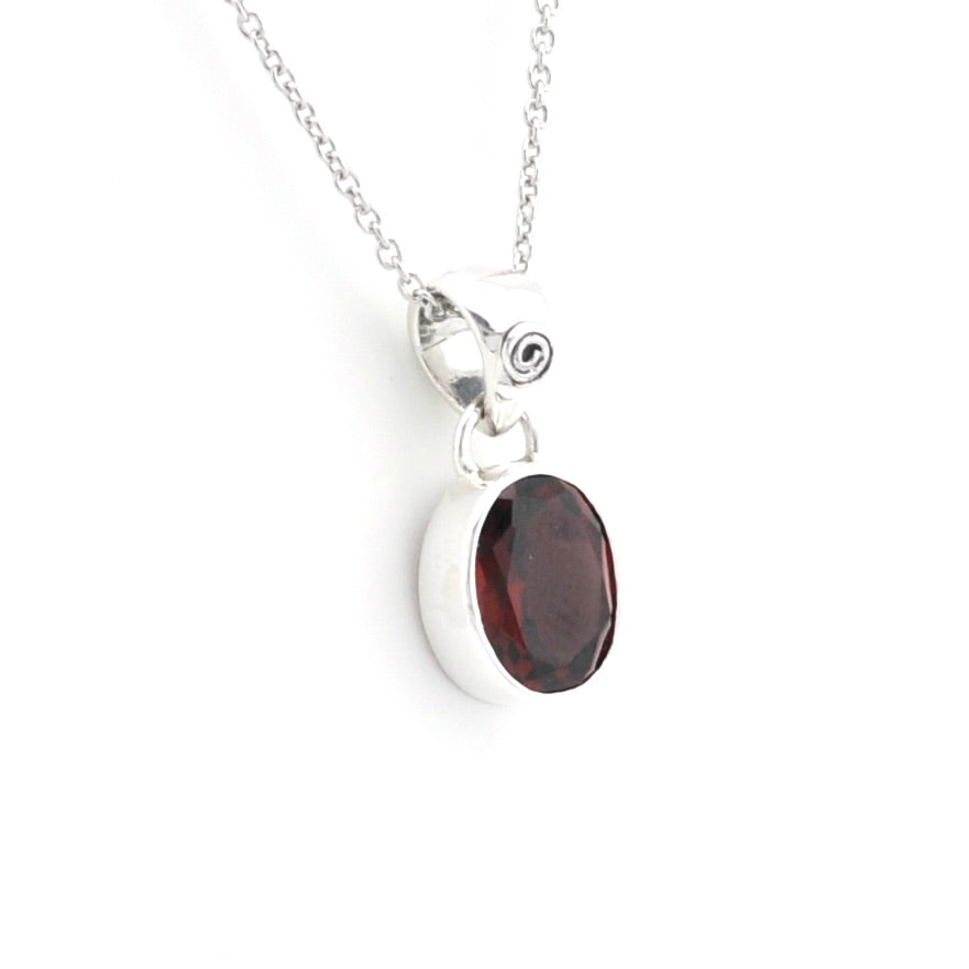 A silver pendant necklace featuring an oval dark red garnet stone in a polished bezel setting, suspended from a delicate silver chain against a plain white background.