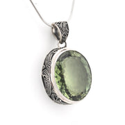 An oval pendant necklace featuring a faceted olive-green gemstone in a silver-toned bezel with ornate details on the sides, suspended from a delicate silver snake chain against a plain white background.