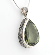 A silver teardrop pendant necklace featuring a faceted olive green gemstone, set in an ornate silver bezel with detailed filigree patterns, suspended from a silver snake chain against a plain white background.
