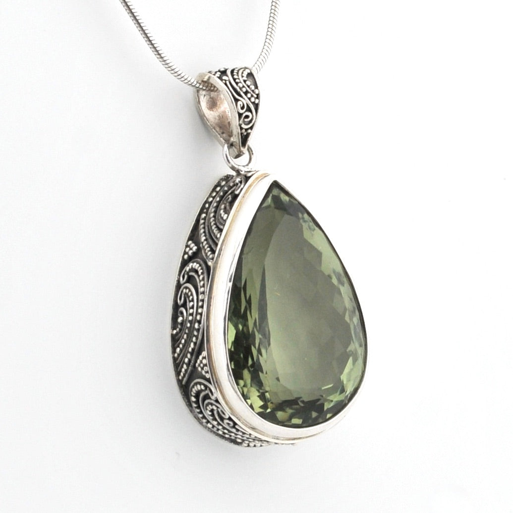 A silver teardrop pendant necklace featuring a faceted olive green gemstone, set in an ornate silver bezel with detailed filigree patterns, suspended from a silver snake chain against a plain white background.