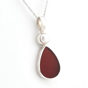 A silver pendant necklace featuring a teardrop-shaped dark reddish-brown sea glass and a small white pearl accent, shown on a white background.