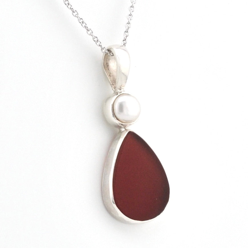A silver pendant necklace featuring a teardrop-shaped dark reddish-brown sea glass and a small white pearl accent, shown on a white background.