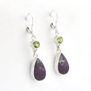 A pair of silver drop earrings featuring a small round green peridot gemstone and a larger teardrop-shaped dark purple stone, displayed on a clean white background.
