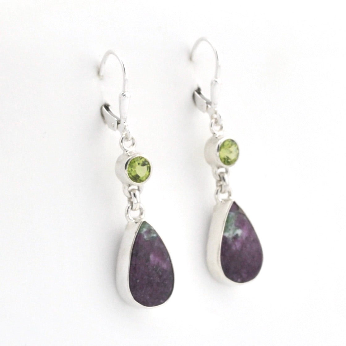 A pair of silver drop earrings featuring a small round green peridot gemstone and a larger teardrop-shaped dark purple stone, displayed on a clean white background.