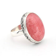 A silver ring featuring a large, oval-shaped pinkish-red stone with a textured, stone-like surface, set in an ornate silver bezel with a polished silver band.
