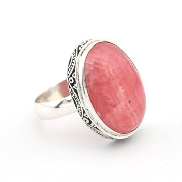 A silver ring featuring a large, oval-shaped pinkish-red stone with a textured, stone-like surface, set in an ornate silver bezel with a polished silver band.