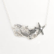 A silver-toned necklace featuring a mermaid tail, swirling wave details, and a starfish pendant with small blue topaz, shown on a plain white background.