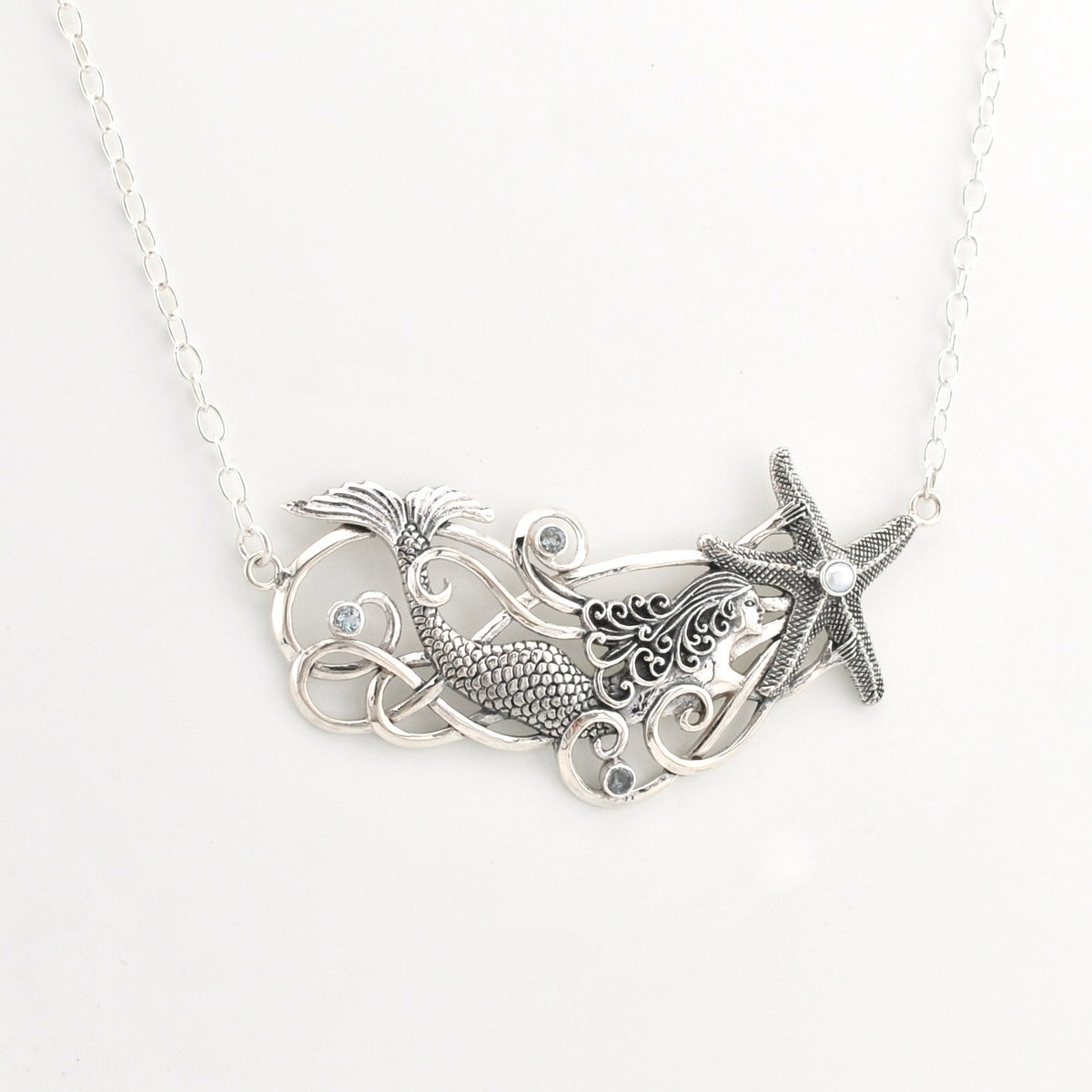 A silver-toned necklace featuring a mermaid tail, swirling wave details, and a starfish pendant with small blue topaz, shown on a plain white background.