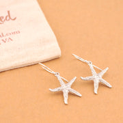 Silver starfish earrings on a beige surface with a jewelry box in the background.