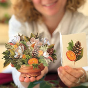 Person holding a small decorative pot with flowers and a card featuring similar design.