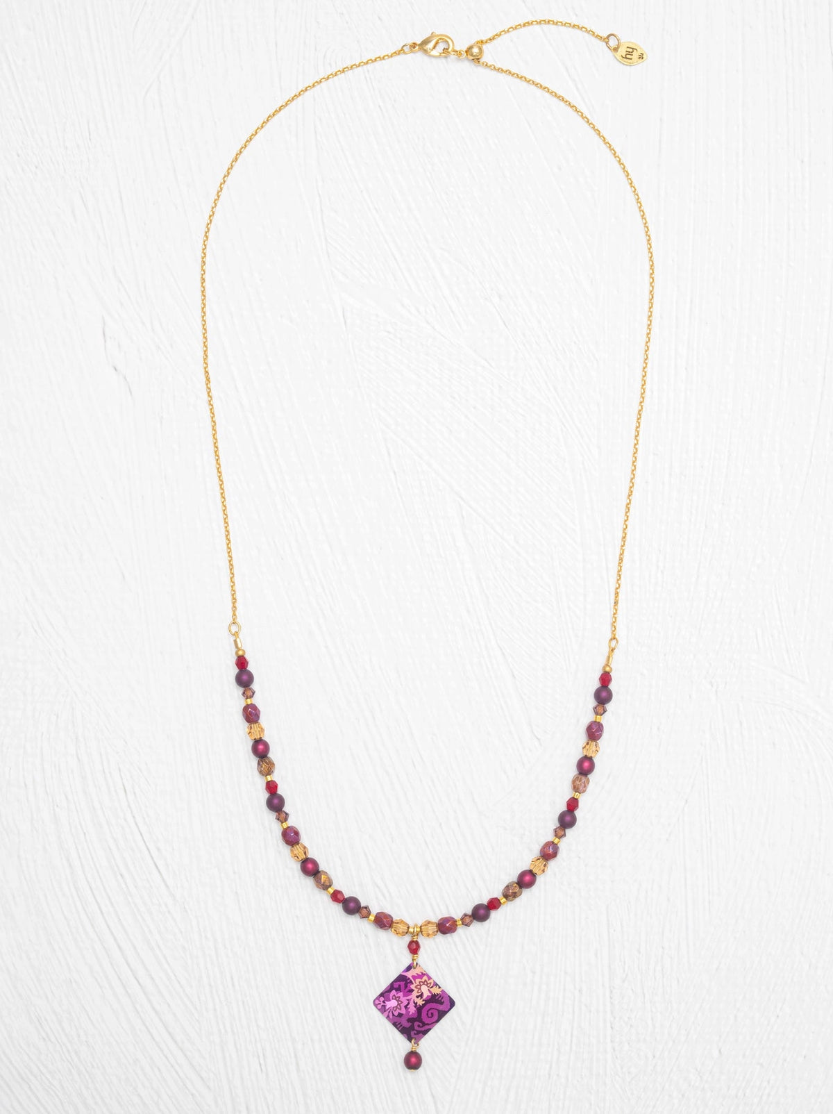 A gold chain necklace featuring a beaded strand with purple and gold beads leading to a purple diamond-shaped pendant, displayed on a white textured background.