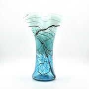 A hand-blown glass vase with a flared rim, featuring a color gradient from light aqua at the top to vibrant blue at the base, decorated with a black branch-like web pattern.