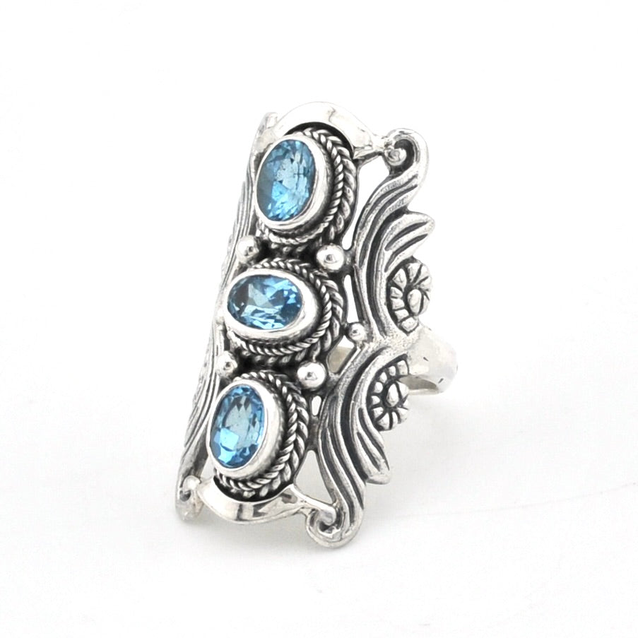 A wide silver-toned ring featuring three vertical oval-cut light blue gemstones in an ornate, textured setting with decorative scrollwork, photographed on a plain white background.