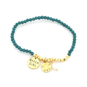 A beaded stretch bracelet with dark teal beads, gold-toned spacer beads, and three charms: a round gold disc stamped 'BEACH GIRL', a gold sand dollar charm, and a small white pearl bead.