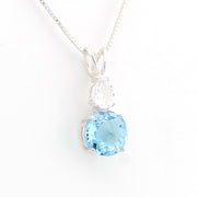 A silver pendant necklace featuring a round light blue gemstone and a smaller marquise-cut clear gemstone, displayed on a white background.