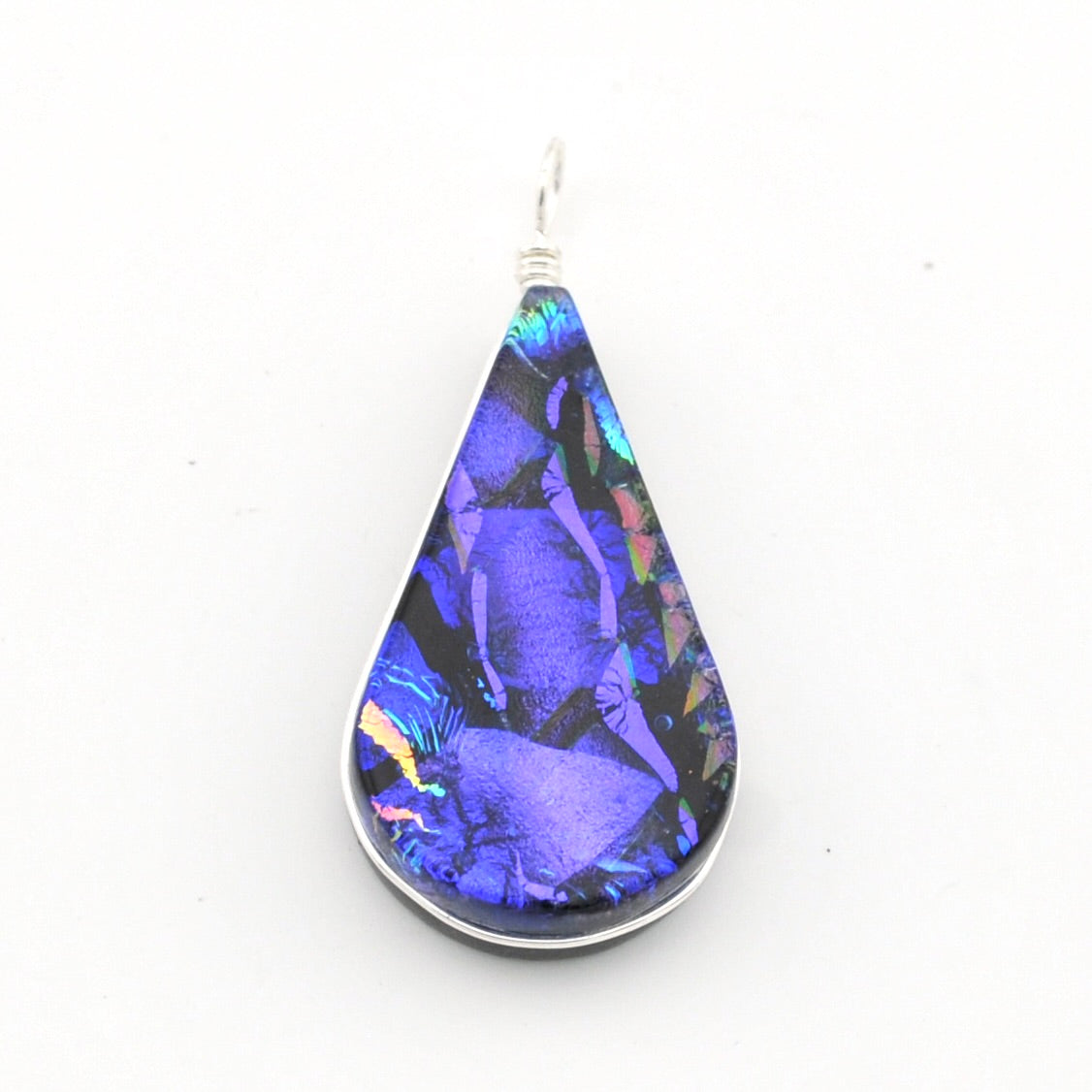 A teardrop-shaped pendant featuring a vibrant blue and purple iridescent dichroic glass set in a silver bezel with a silver wire-wrapped bail, shown on a plain white background.