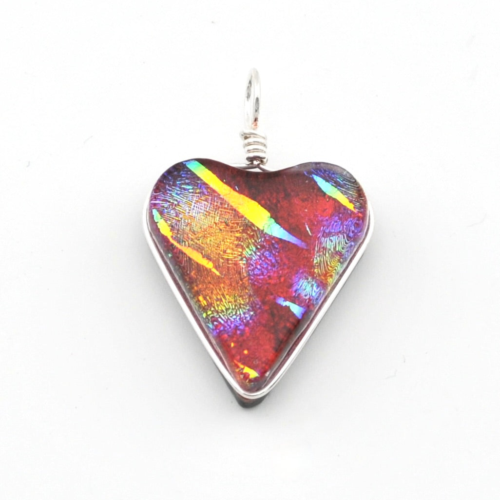 A heart-shaped glass pendant featuring a vibrant, iridescent play-of-color pattern with red, orange, yellow, and blue hues, set in a silver-colored metal bezel with a silver wire-wrapped bail on a white background.