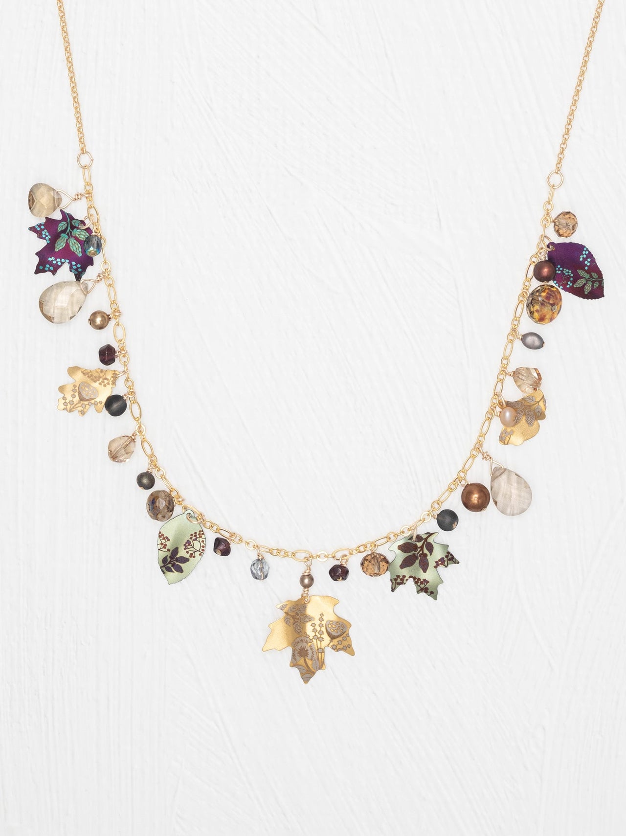 A gold-tone statement necklace featuring multiple dangling charms, including enamel maple leaves in green, purple, and gold, clear teardrop beads, and small dark and metallic beads, displayed against a textured white background.