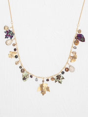 A gold-tone statement necklace featuring multiple dangling charms, including enamel maple leaves in green, purple, and gold, clear teardrop beads, and small dark and metallic beads, displayed against a textured white background.