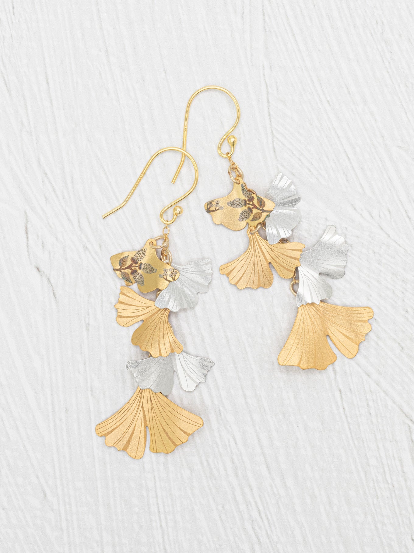 A pair of gold and silver dangling earrings with a fan-shaped leaf design, displayed on a light gray textured background.