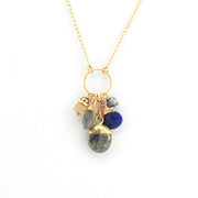 A gold chain necklace with a circular ring pendant holding several small gemstone charms, including a dark gray labradorite, a deep blue stone, and a pale peach stone, shown on a white background.