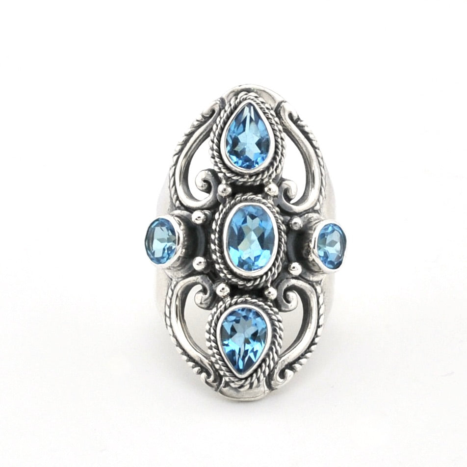 A silver-toned ring featuring five light blue gemstones in an ornate filigree design, set against a plain white background.
