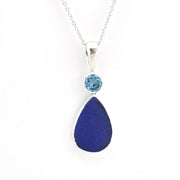 A silver pendant necklace featuring a teardrop-shaped dark blue sea glass drop and a small round light blue gemstone above it, set on a delicate silver chain against a white background.