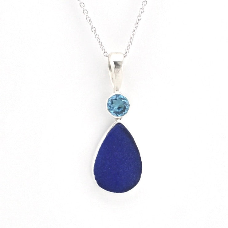 A silver pendant necklace featuring a teardrop-shaped dark blue sea glass drop and a small round light blue gemstone above it, set on a delicate silver chain against a white background.