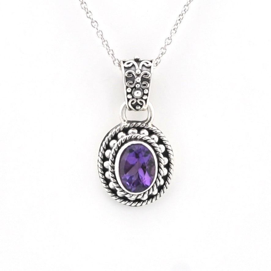 A silver-toned oval pendant necklace featuring a central purple amethyst gemstone, detailed with textured rope-like borders and a decorative filigree bail, suspended from a delicate silver chain against a plain white background.